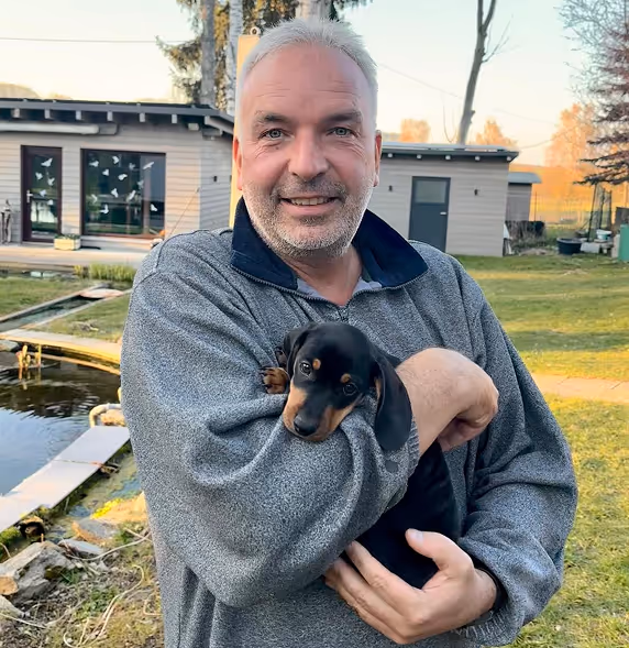 Ein lächelnder Mann hält einen kleinen schwarz-braunen Welpen im Arm, im Hintergrund ein Garten mit Gebäude und Teich.